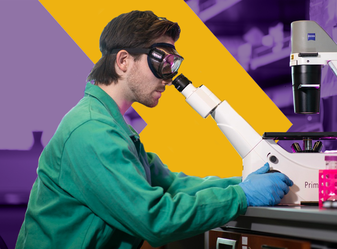 A student wearing a lab coat and goggles looking through a microscope.