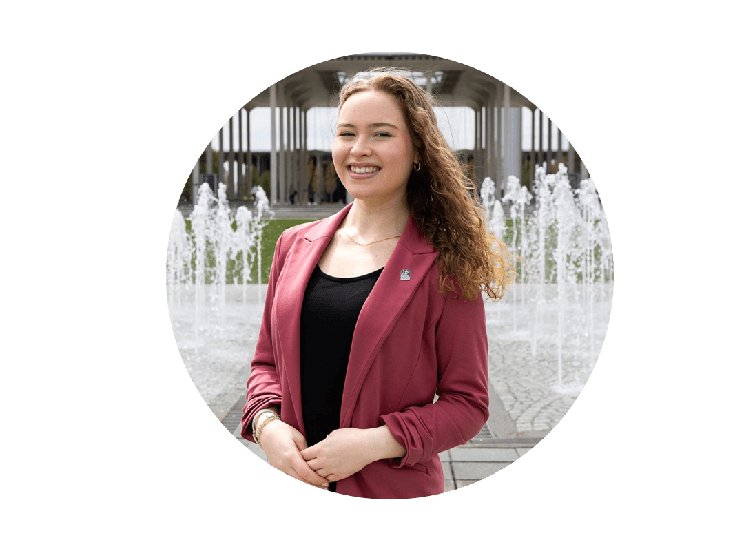 A student wearing a maroon blazer standing in front of a water fountain.
